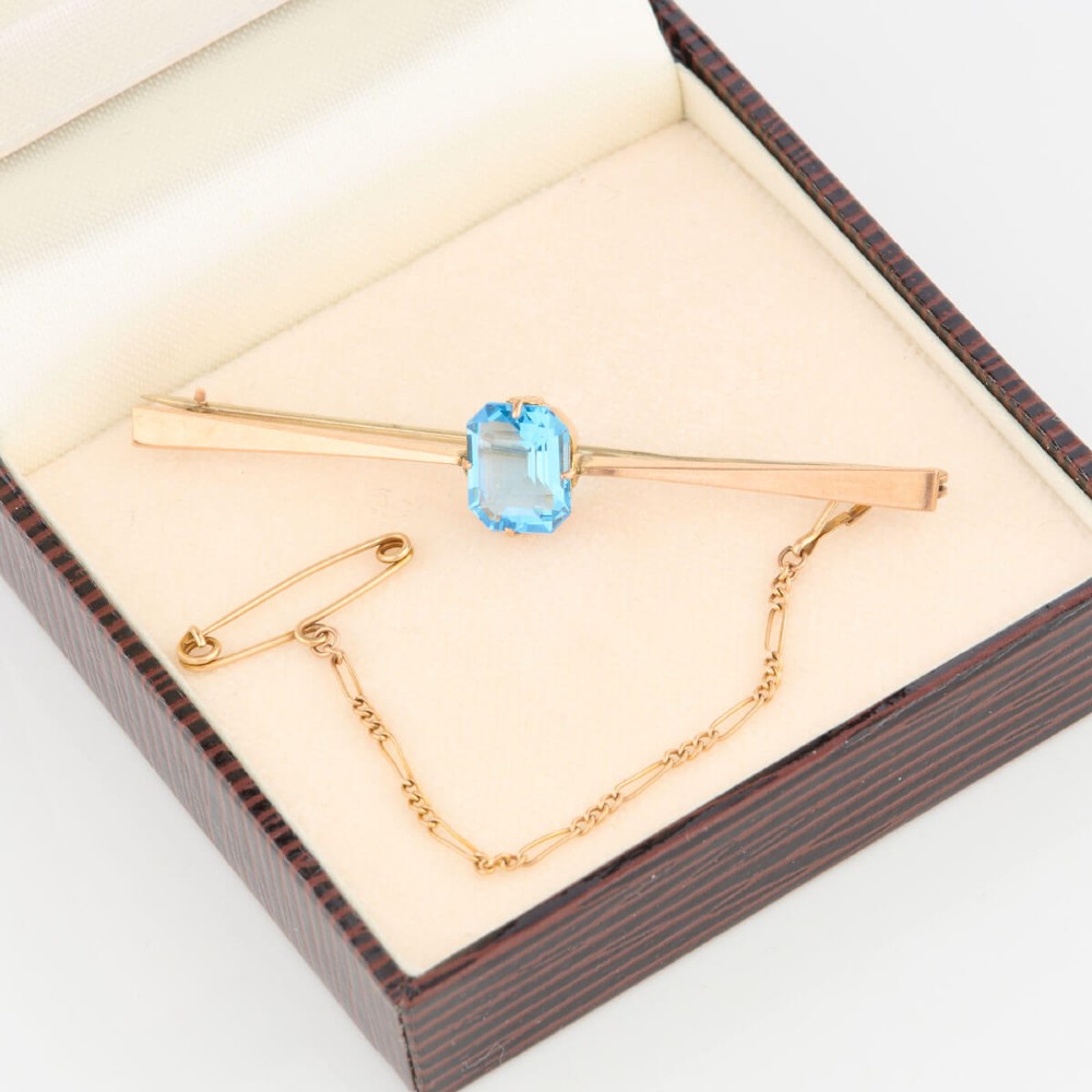 A 9ct yellow gold horizontal bar brooch with a central emerald-cut blue paste stone and an attached safety chain. View VII