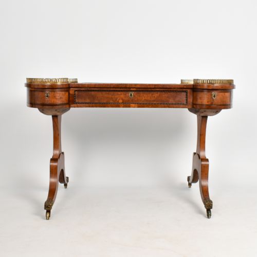 fig 1. A Regency period burr oak writing desk featuring a green leather inset top, brass gallery rails on raised side tiers, a central drawer, and trestle legs on brass castors.