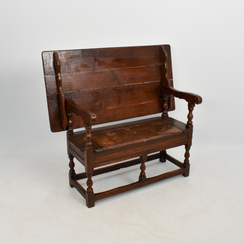 An 18th-century dark oak monks bench featuring a convertible tilt-top backrest, turned arm supports, and block legs with perimeter stretchers.