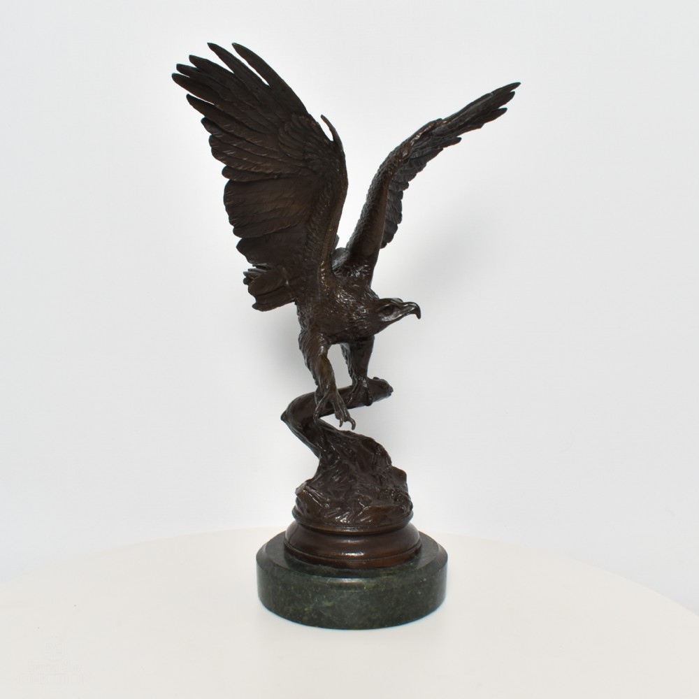 A dark brown bronze sculpture depicting an eagle with wings raised perched on a branch, mounted on a round green marble plinth.