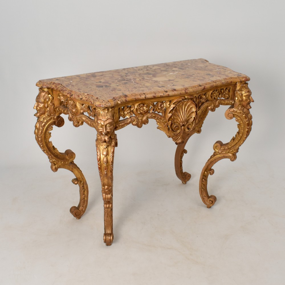 A 19th-century giltwood console table featuring carved grotesque masks, central shell carving, and a pink and beige marble top.