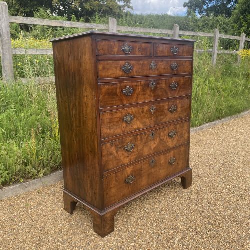 fig 1. A tall George II walnut chest of drawers featuring figured veneer drawer fronts, brass keyhole escutcheons, brass handles, and bracket feet.