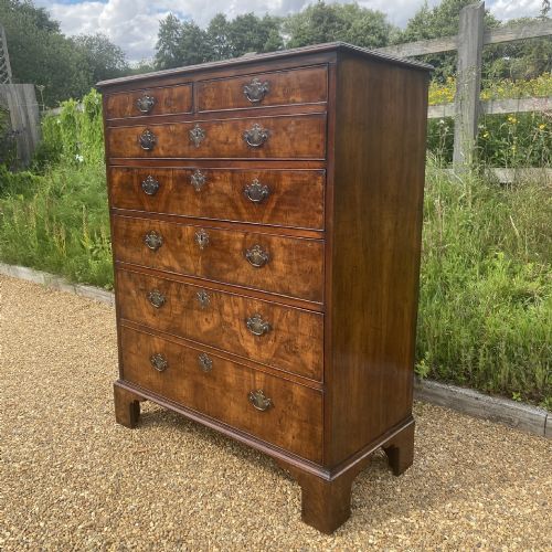 fig 2. A tall George II walnut chest of drawers featuring figured veneer drawer fronts, brass keyhole escutcheons, brass handles, and bracket feet.