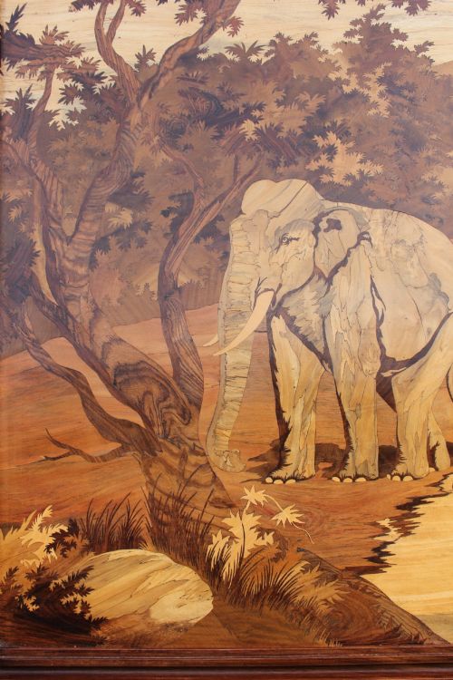 fig 1. A horizontal framed wooden marquetry panel depicting a herd of elephants bathing in a river surrounded by trees and mountains made from contrasting timber veneers.