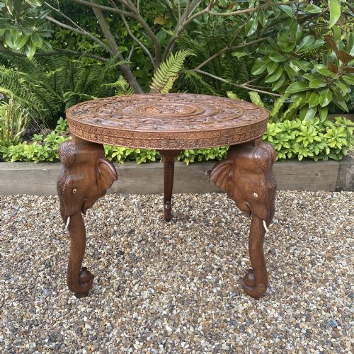 fig 1. A circular carved rosewood side table supported by three legs shaped as detailed elephant heads with trunks extending to the floor.