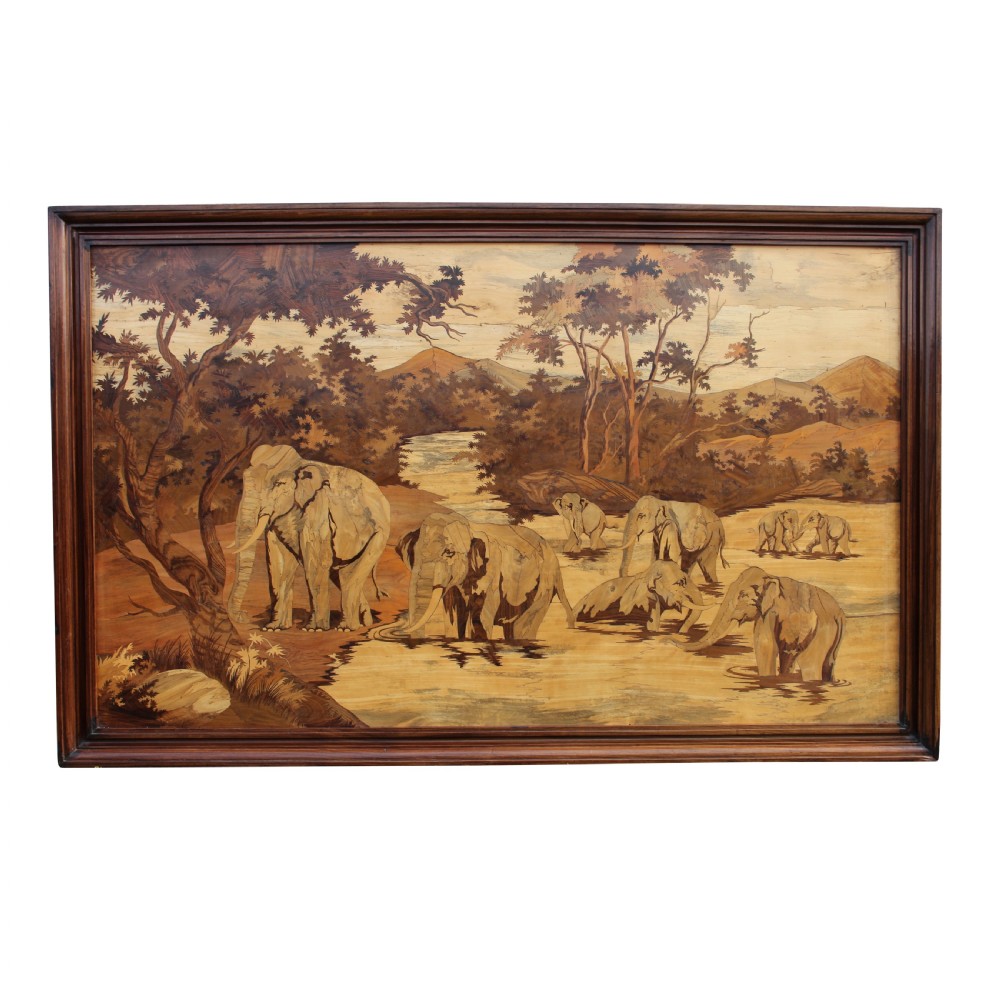 A large rectangular wooden marquetry panel depicting a herd of elephants bathing in a river surrounded by trees and mountains inside a moulded dark wood frame.