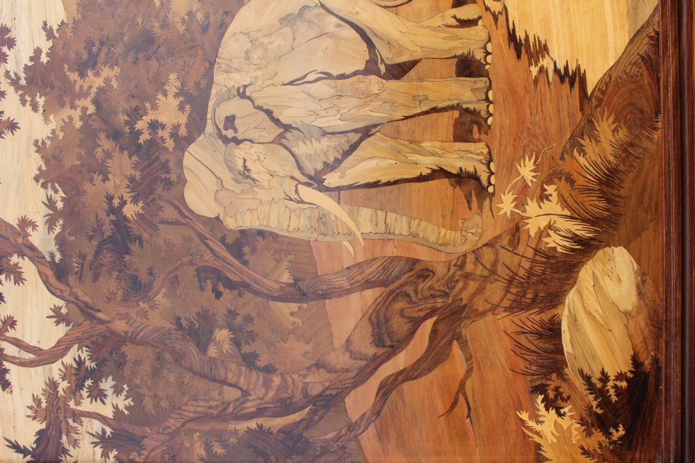 A large rectangular wooden marquetry panel depicting a herd of elephants bathing in a river surrounded by trees and mountains inside a moulded dark wood frame. View II