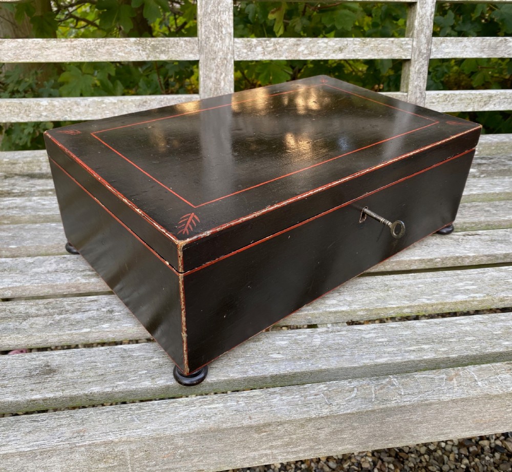 A dark painted rectangular wooden box featuring red line borders, corner leaf motifs, turned feet, and a key in the front lock.