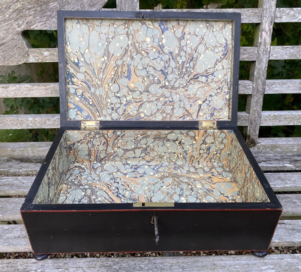 A dark painted rectangular wooden box featuring red line borders, corner leaf motifs, turned feet, and a key in the front lock. View III
