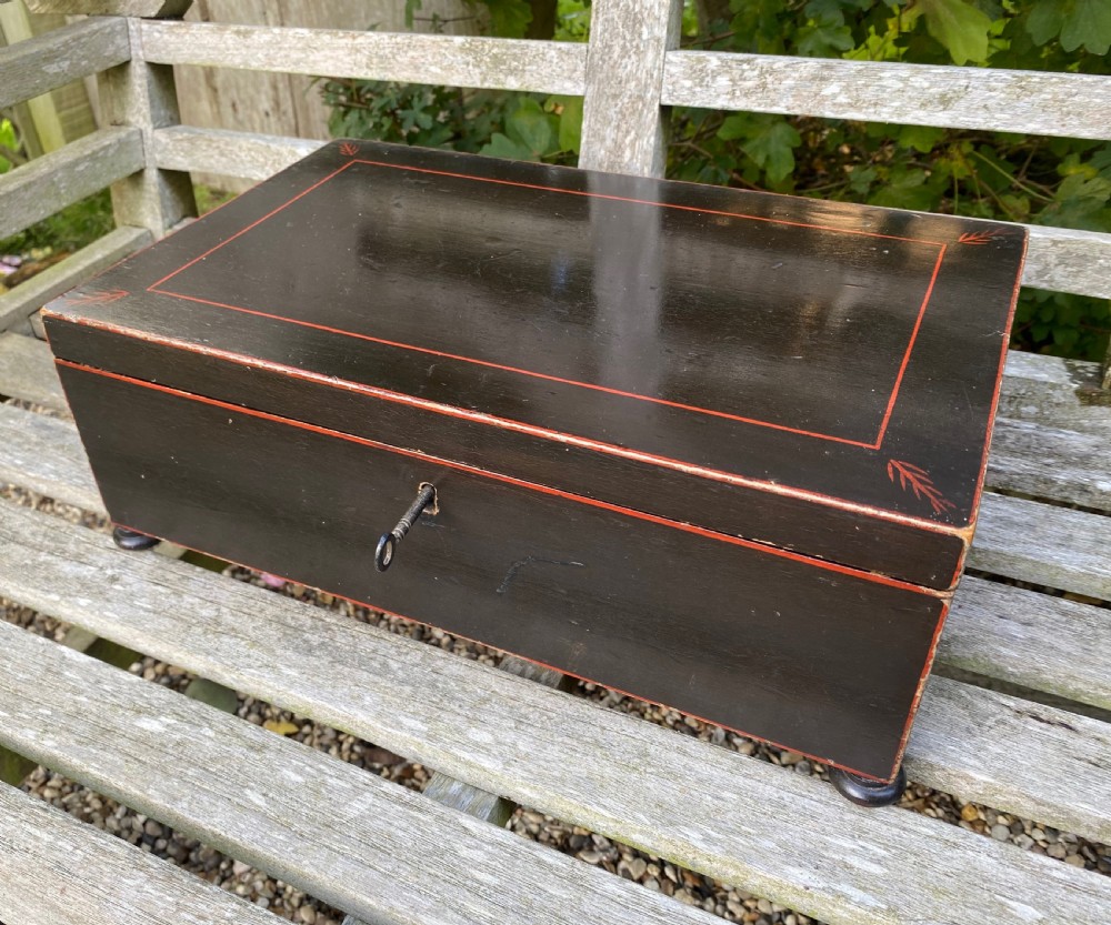 A dark painted rectangular wooden box featuring red line borders, corner leaf motifs, turned feet, and a key in the front lock. View IV