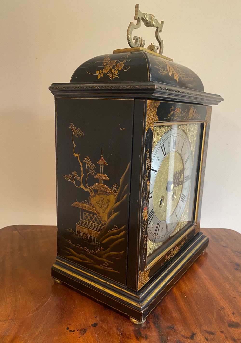 A black lacquered bracket clock decorated with gold gilt Chinoiserie landscapes, featuring a brass carrying handle on top and a silvered dial ring with Roman numerals. View II