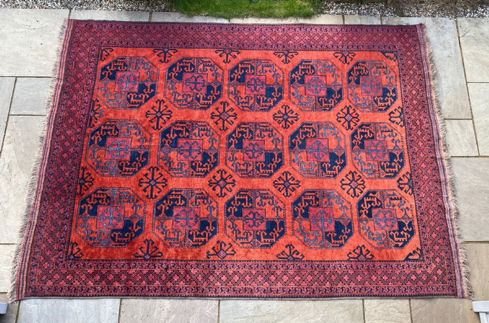 A hand-woven antique Turkmen wool rug featuring a repeating pattern of dark blue octagonal gol motifs on a terracotta red field with a geometric border.