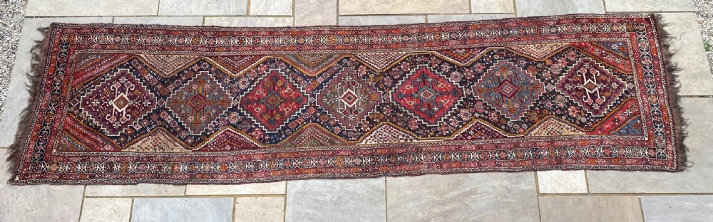 An antique Persian Shiraz wool runner rug featuring a sequence of geometric diamond medallions in crimson red and navy blue with fringed ends.