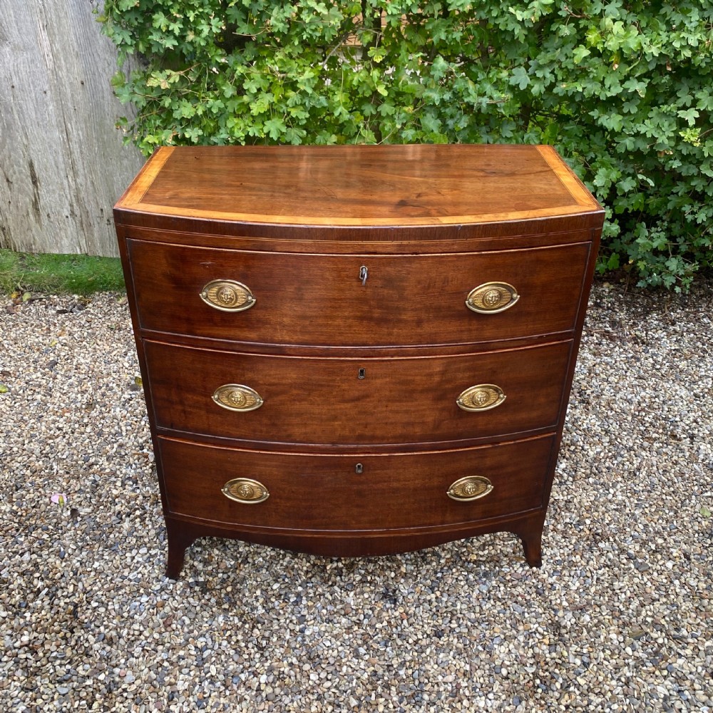 An early nineteenth-century mahogany bow-fronted chest of drawers featuring satinwood crossbanding, three graduated drawers, and oval brass handles.