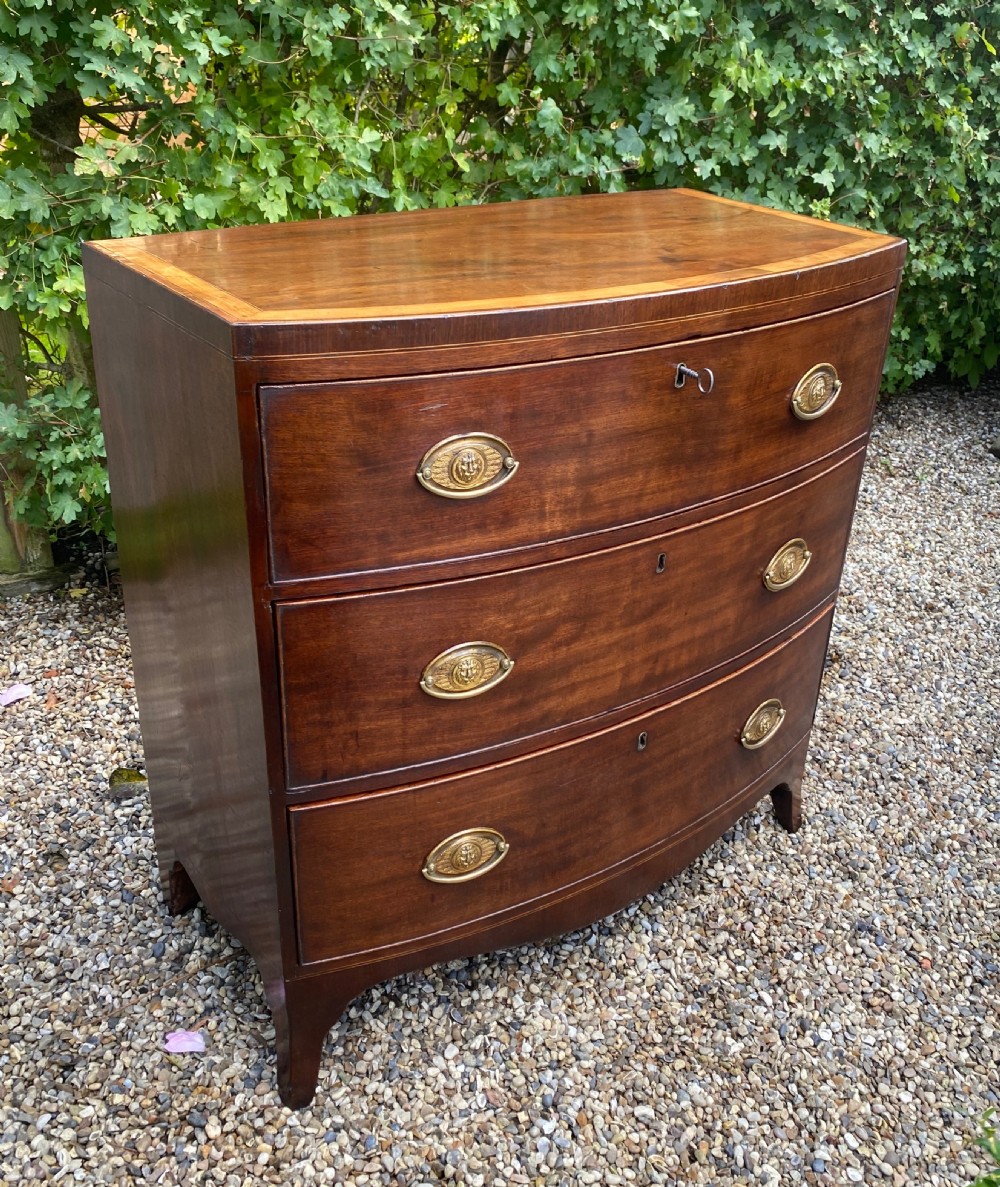 An early nineteenth-century mahogany bow-fronted chest of drawers featuring satinwood crossbanding, three graduated drawers, and oval brass handles. View V