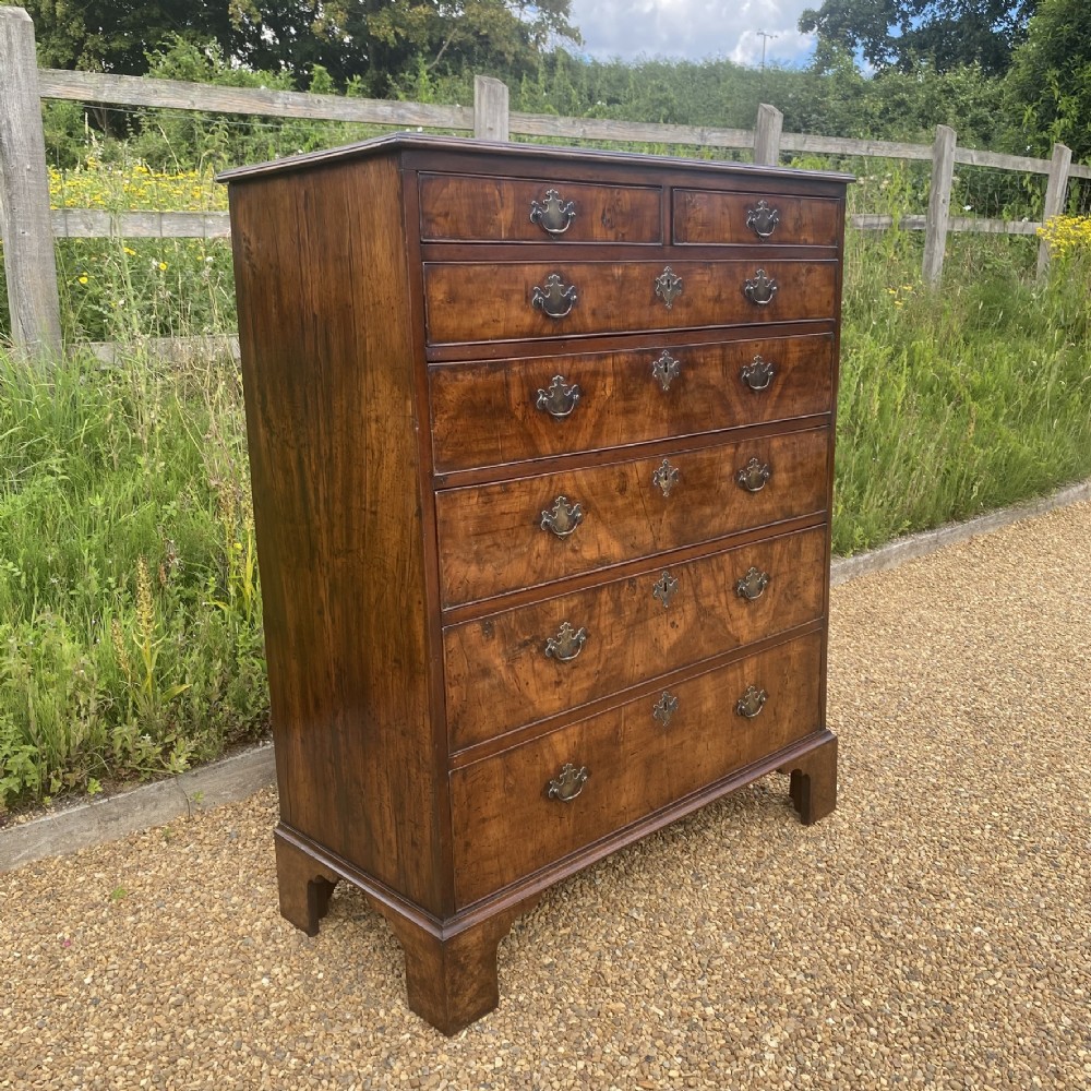 A tall George II walnut chest of drawers featuring figured veneer drawer fronts, brass keyhole escutcheons, brass handles, and bracket feet. View II