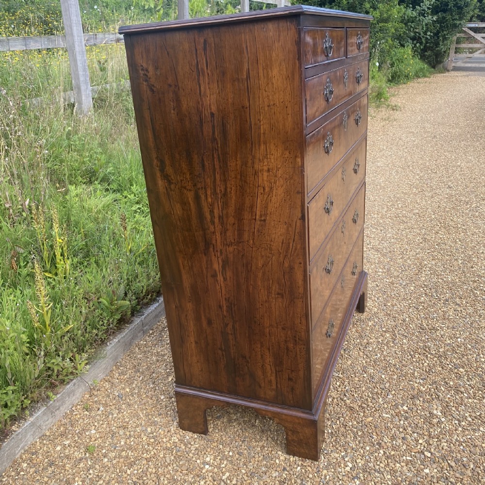 A tall George II walnut chest of drawers featuring figured veneer drawer fronts, brass keyhole escutcheons, brass handles, and bracket feet. View IX