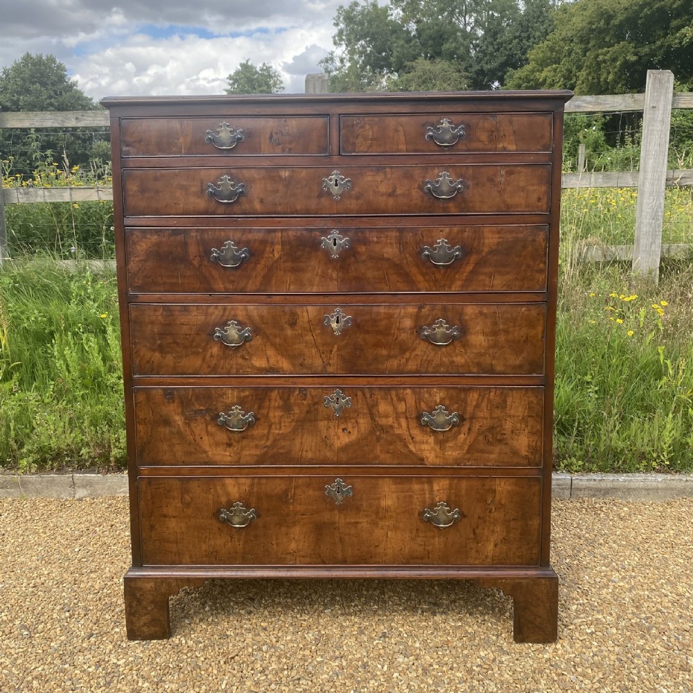 A tall George II walnut chest of drawers featuring figured veneer drawer fronts, brass keyhole escutcheons, brass handles, and bracket feet. View X