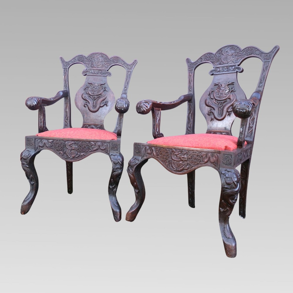 A pair of dark carved hardwood armchairs featuring high back splats with dragon relief motifs, dragon head terminal arms, and red upholstered seats.
