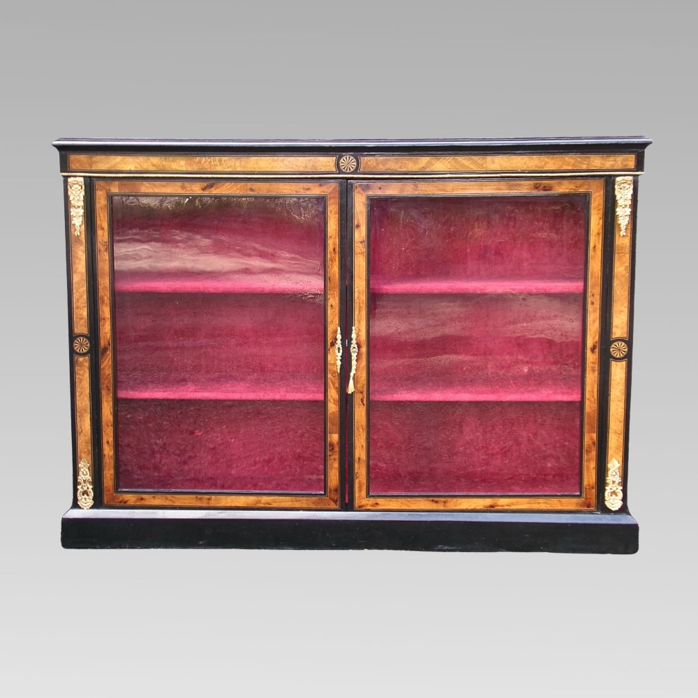 A 19th-century Victorian walnut and ebonised display cabinet featuring two glazed doors, gilt metal mounts, and a magenta velvet interior.