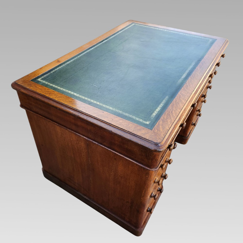 A Victorian oak pedestal writing desk featuring a green leather inset top and multiple drawers with turned wooden handles. View IV
