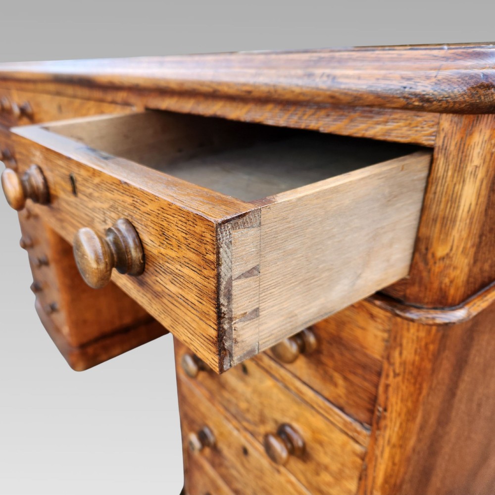A Victorian oak pedestal writing desk featuring a green leather inset top and multiple drawers with turned wooden handles. View VI