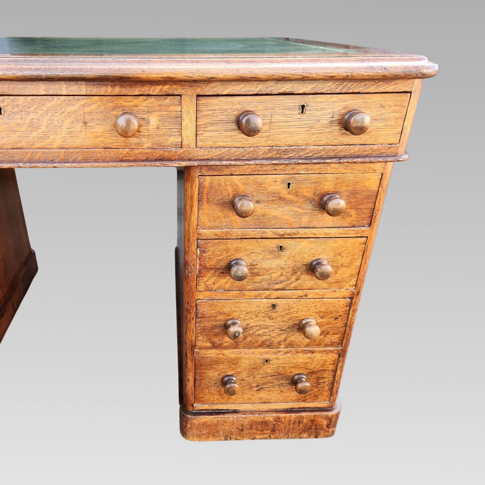 A Victorian oak pedestal writing desk featuring a green leather inset top and multiple drawers with turned wooden handles. View X