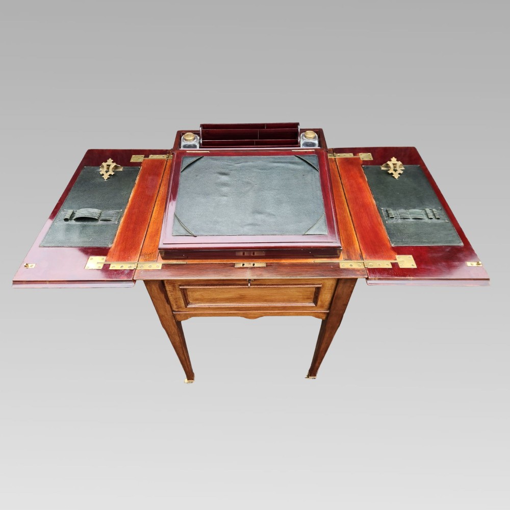 An open antique Edwardian mahogany metamorphic writing desk featuring fold-out side flaps, a green leather writing surface, integrated inkwells, and brass fittings.