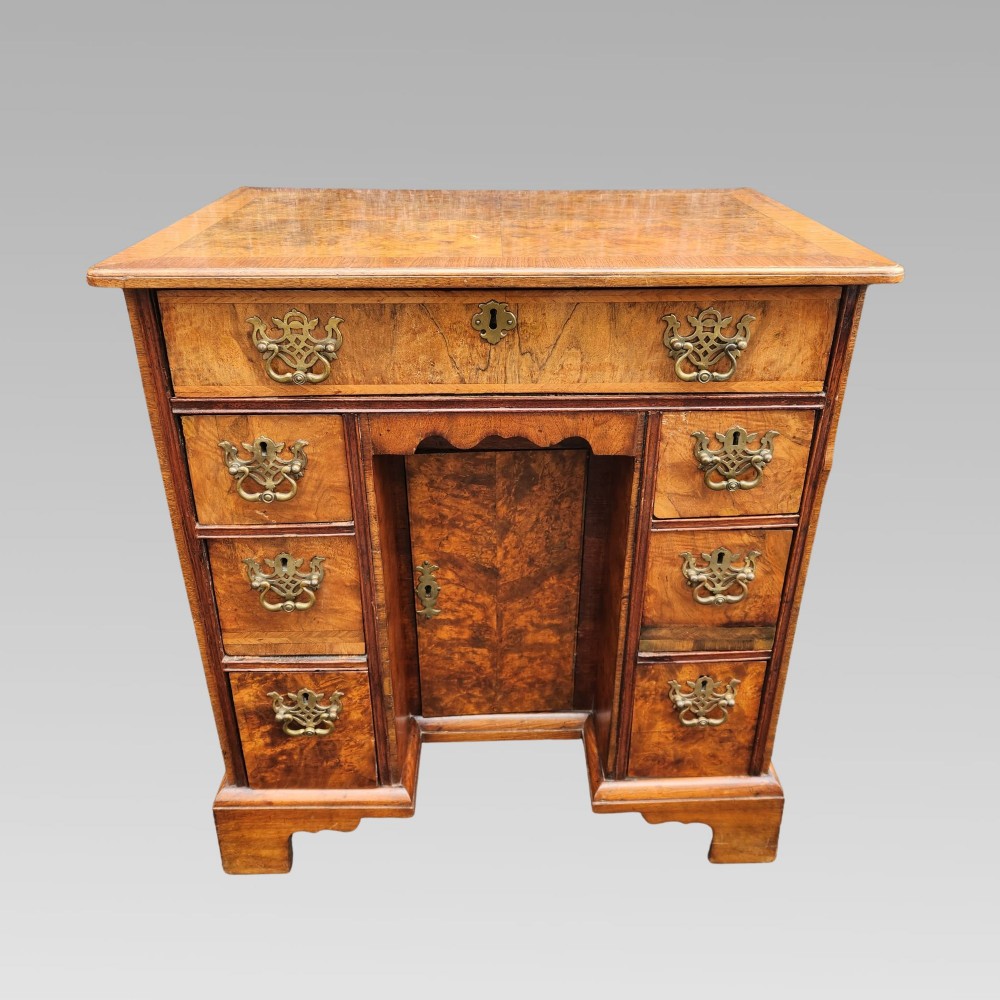 An antique Queen Anne style burr walnut kneehole desk featuring a crossbanded top, brass drop handles, a central recessed cupboard, and bracket feet.