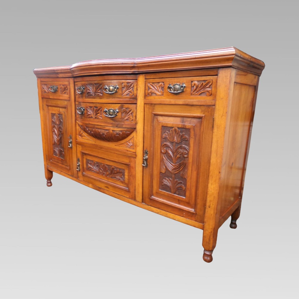 A carved Edwardian mahogany sideboard featuring a bow-front centre section with drawers, flanked by cupboard doors with stylised plant motifs. View IV