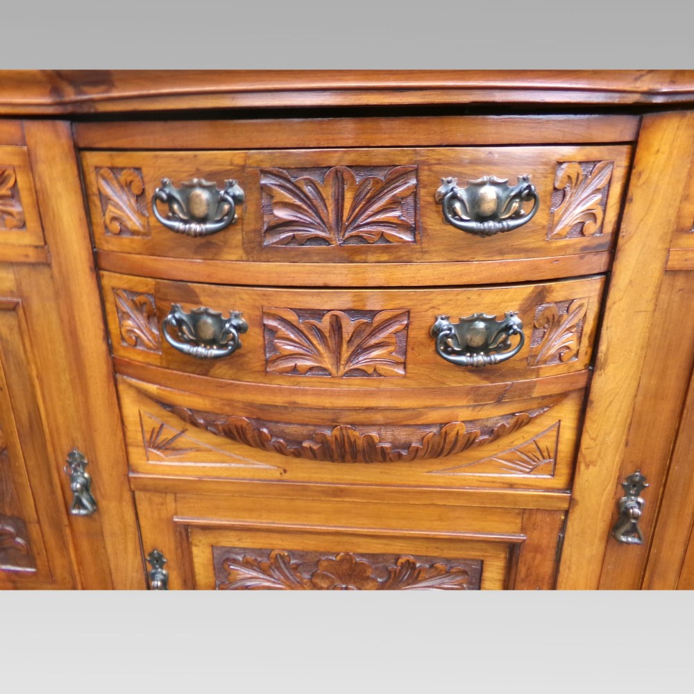 A carved Edwardian mahogany sideboard featuring a bow-front centre section with drawers, flanked by cupboard doors with stylised plant motifs. View VI