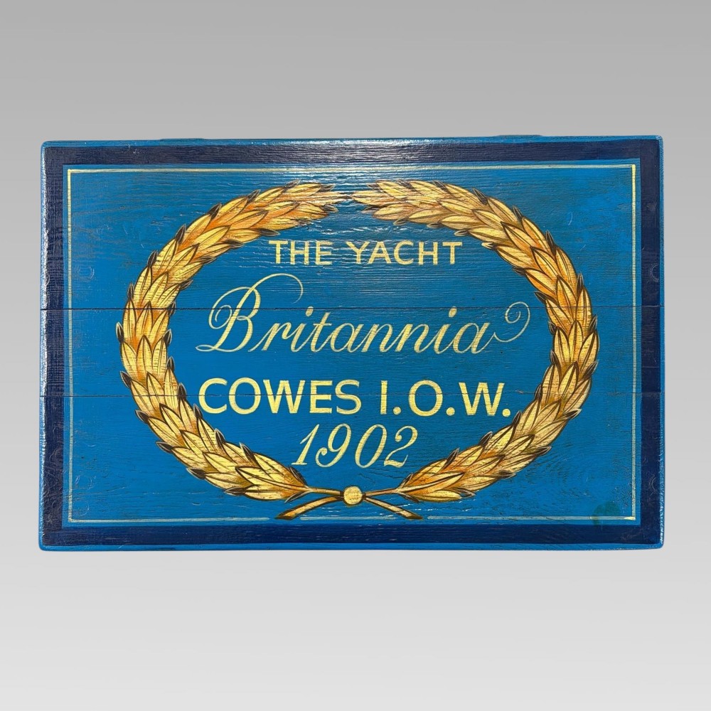 A blue painted wooden storage box featuring a hand-painted scene of sailing yachts and lettering reading The Yacht Britannia Cowes I.O.W. 1902. View II