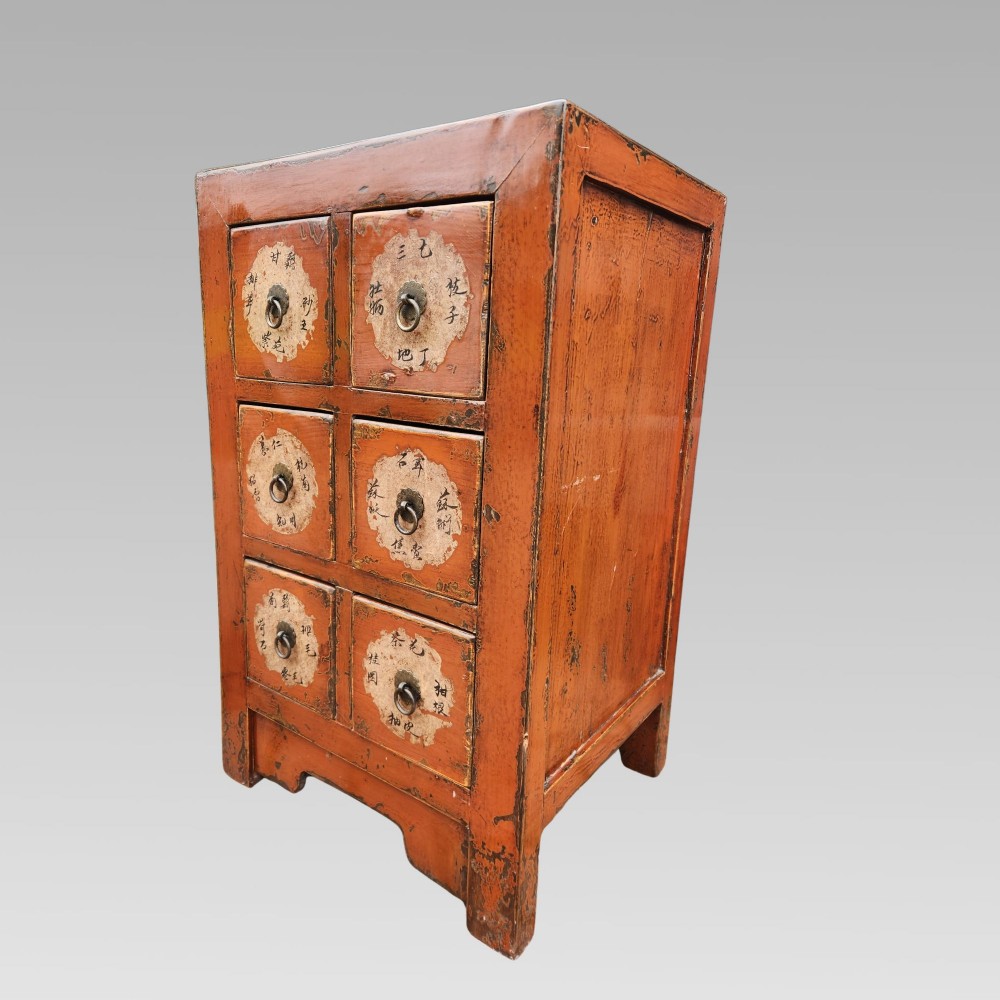 A terracotta red lacquered wooden Chinese apothecary cabinet featuring six drawers with paper calligraphy labels and central metal ring pulls.