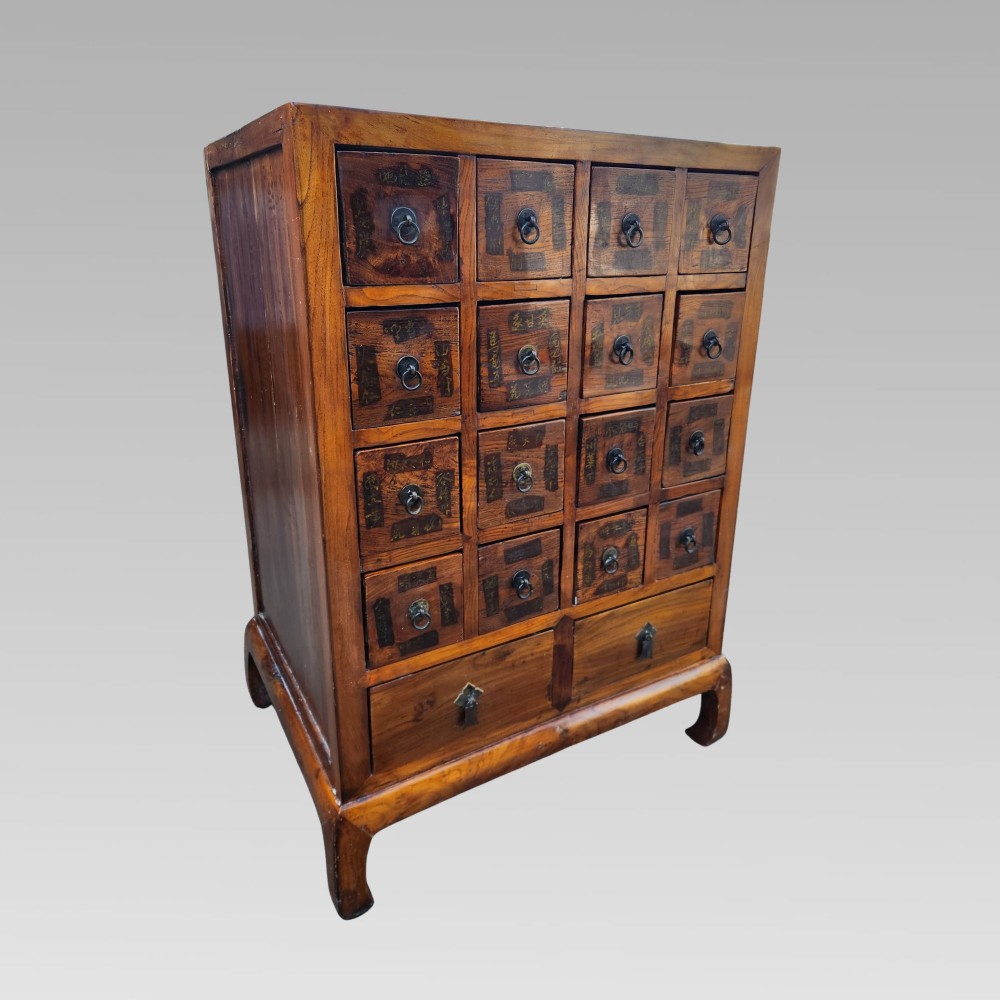A tall warm brown elm apothecary chest featuring sixteen small labelled drawers above two wider base drawers with metal ring pulls. View II