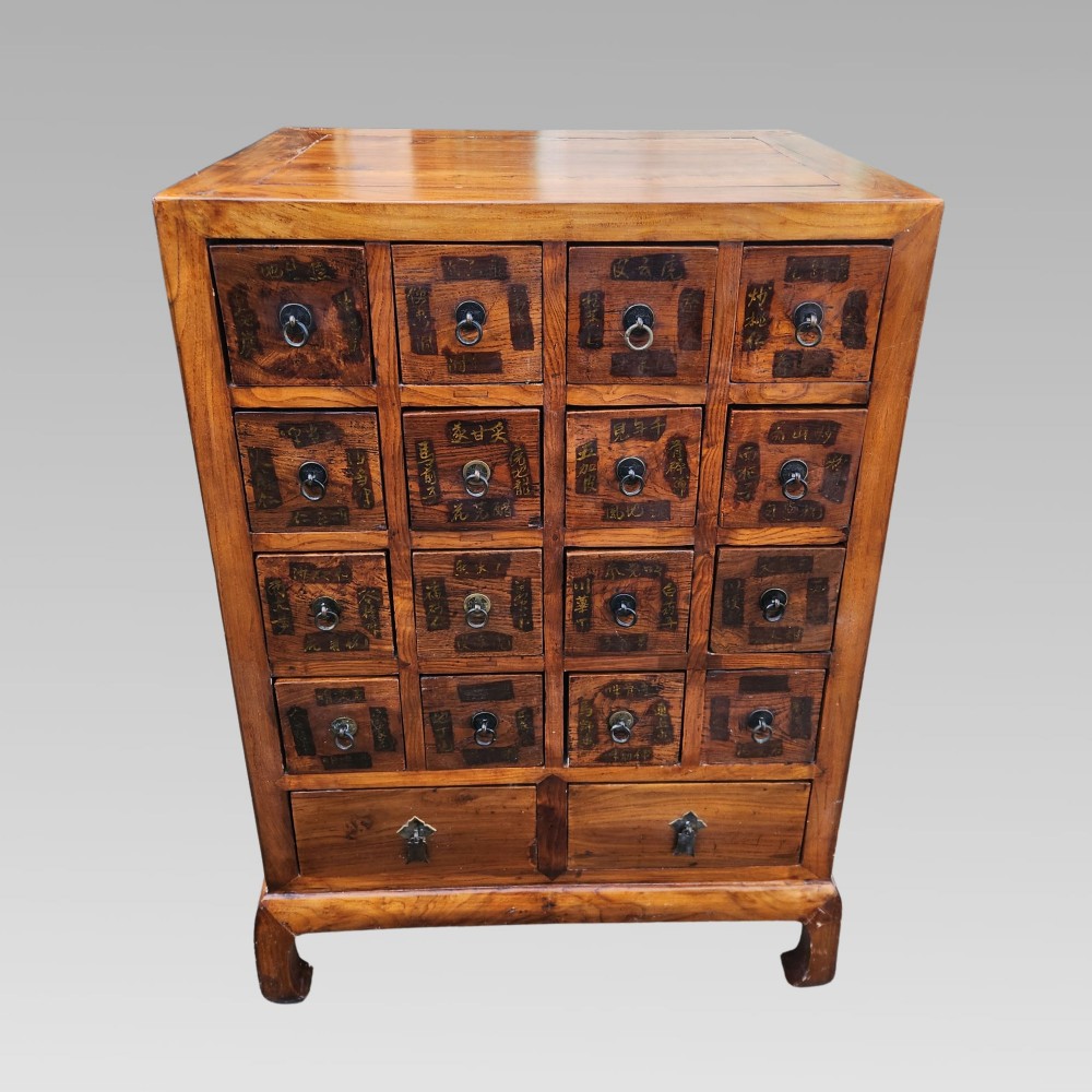 A tall warm brown elm apothecary chest featuring sixteen small labelled drawers above two wider base drawers with metal ring pulls. View IV