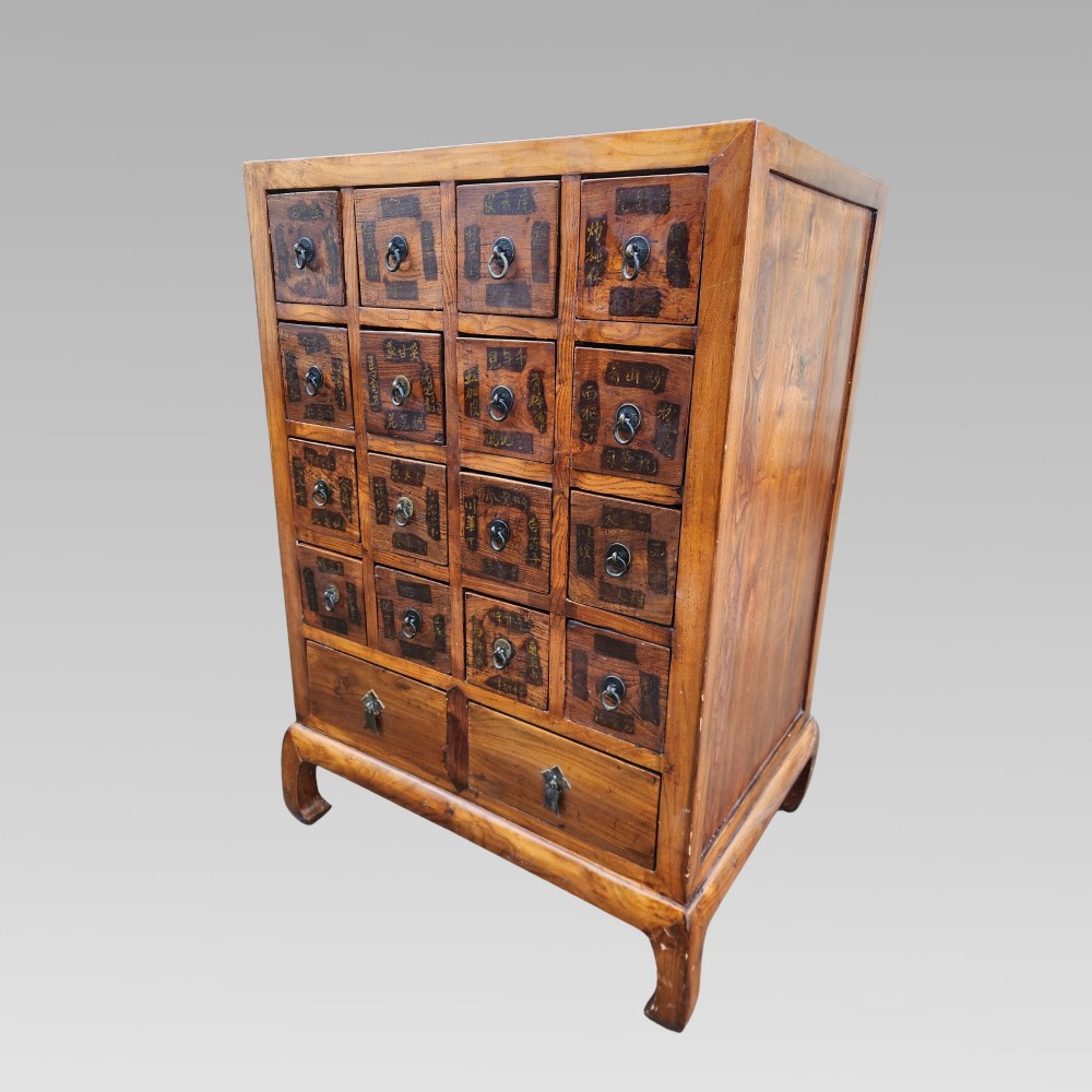 A tall warm brown elm apothecary chest featuring sixteen small labelled drawers above two wider base drawers with metal ring pulls.