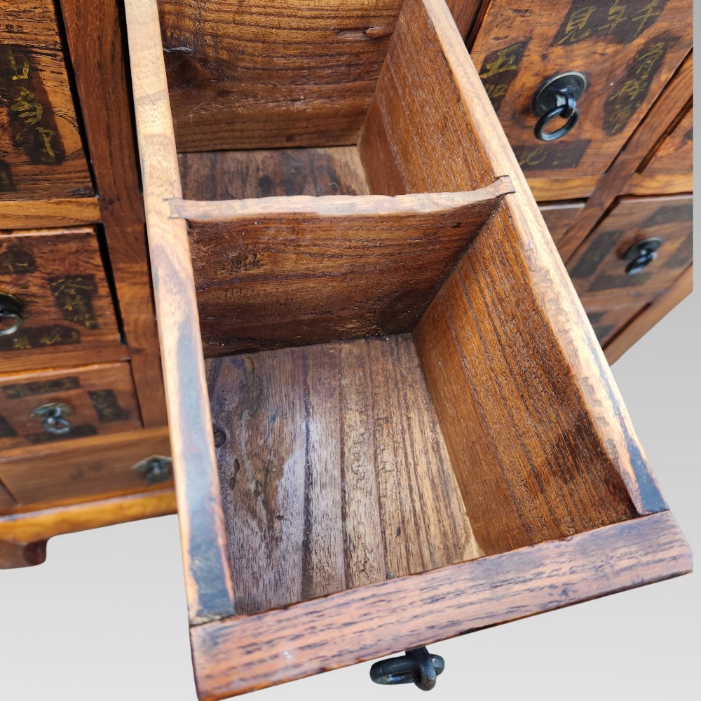 A tall warm brown elm apothecary chest featuring sixteen small labelled drawers above two wider base drawers with metal ring pulls. View XI