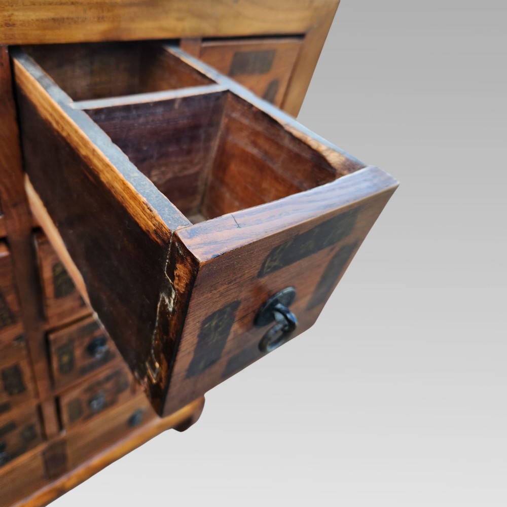 A tall warm brown elm apothecary chest featuring sixteen small labelled drawers above two wider base drawers with metal ring pulls. View XII