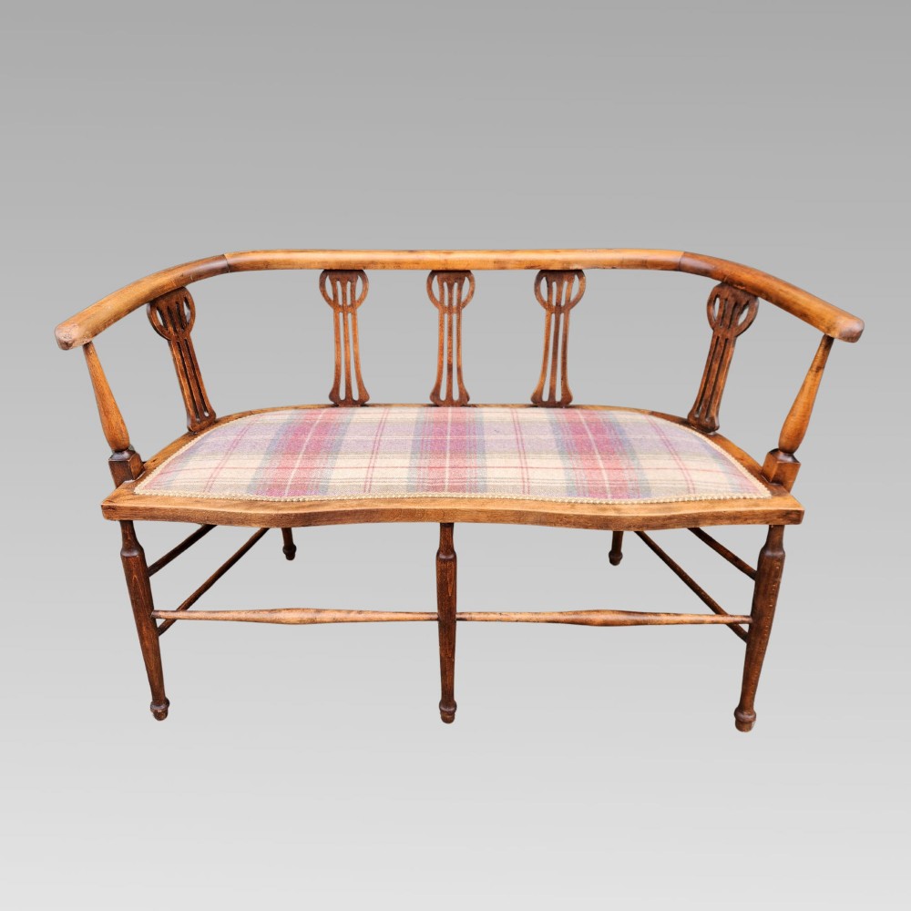 An Edwardian wooden salon settee featuring a curved armrail, pierced splats, turned legs, and a pastel tartan upholstered seat.