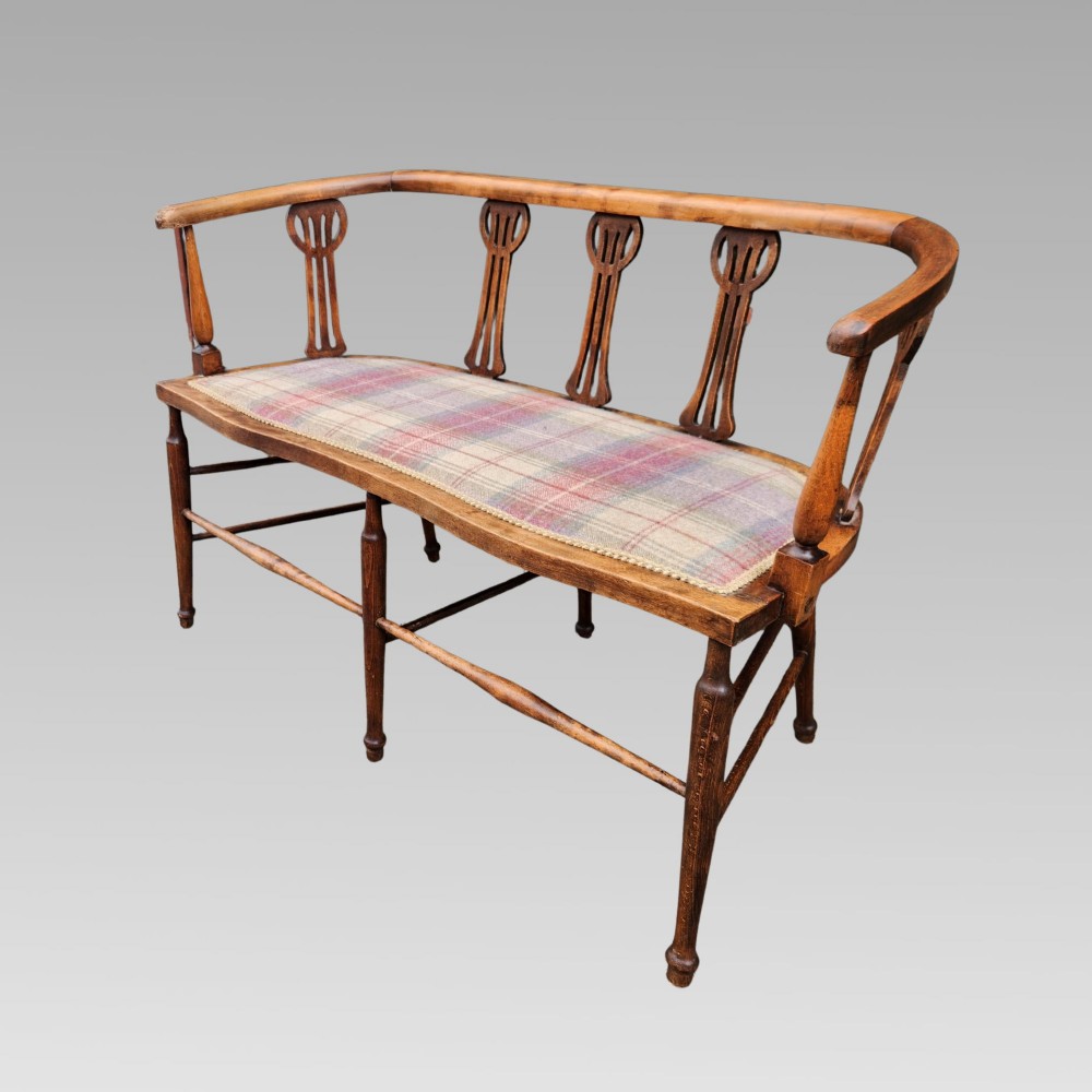 An Edwardian wooden salon settee featuring a curved armrail, pierced splats, turned legs, and a pastel tartan upholstered seat. View V