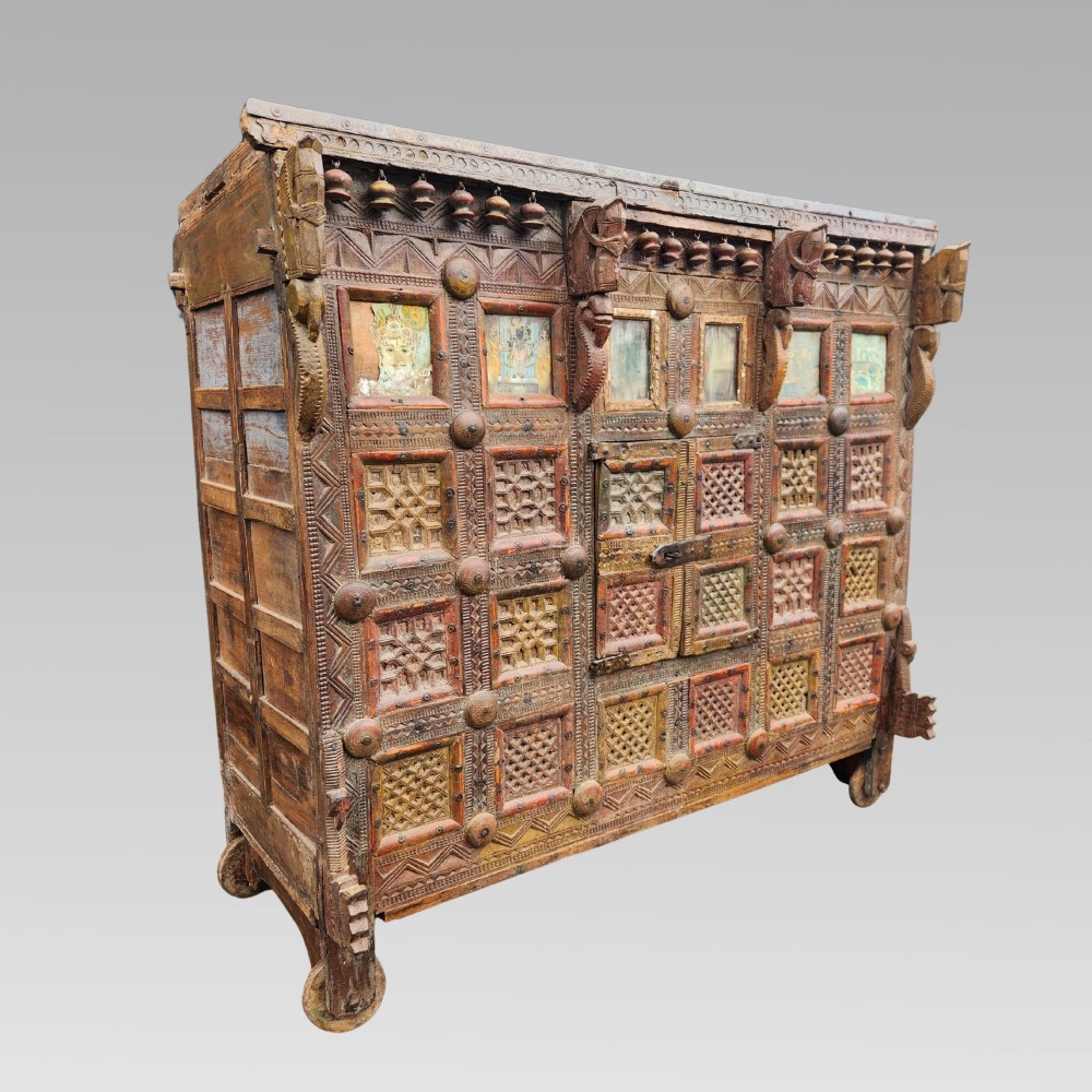 An intricately carved antique Indian teak Damchiya dowry chest featuring painted glass panels, horse head bracket carvings, metal studs, and small wooden wheels.