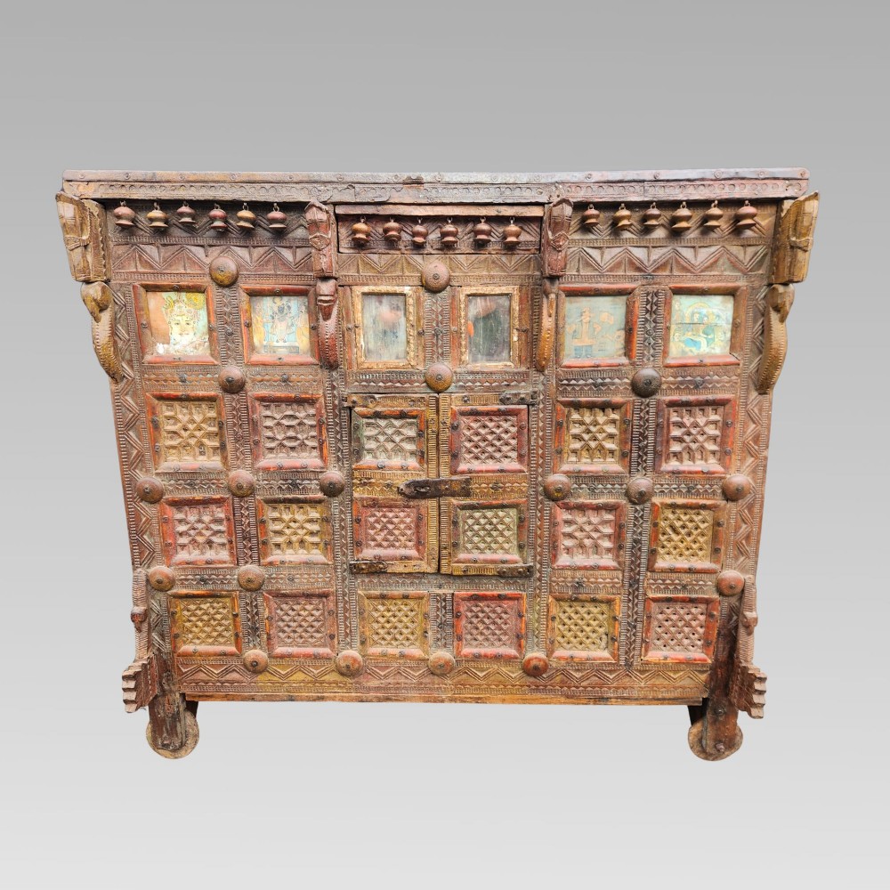 An intricately carved antique Indian teak Damchiya dowry chest featuring painted glass panels, horse head bracket carvings, metal studs, and small wooden wheels. View II