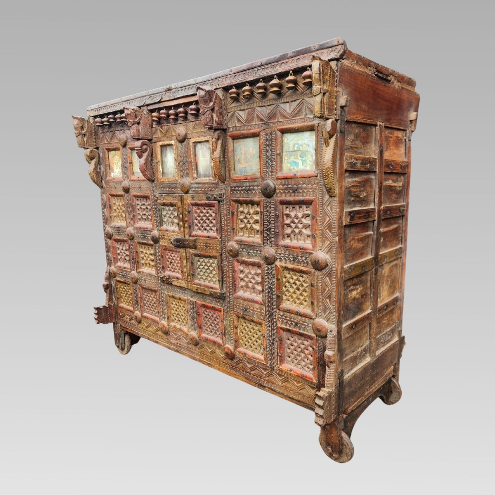An intricately carved antique Indian teak Damchiya dowry chest featuring painted glass panels, horse head bracket carvings, metal studs, and small wooden wheels. View III