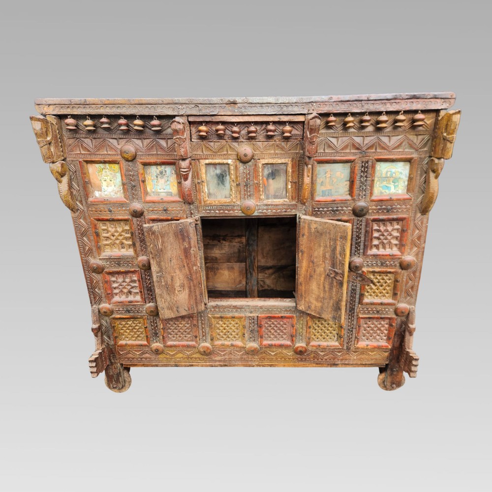 An intricately carved antique Indian teak Damchiya dowry chest featuring painted glass panels, horse head bracket carvings, metal studs, and small wooden wheels. View IV