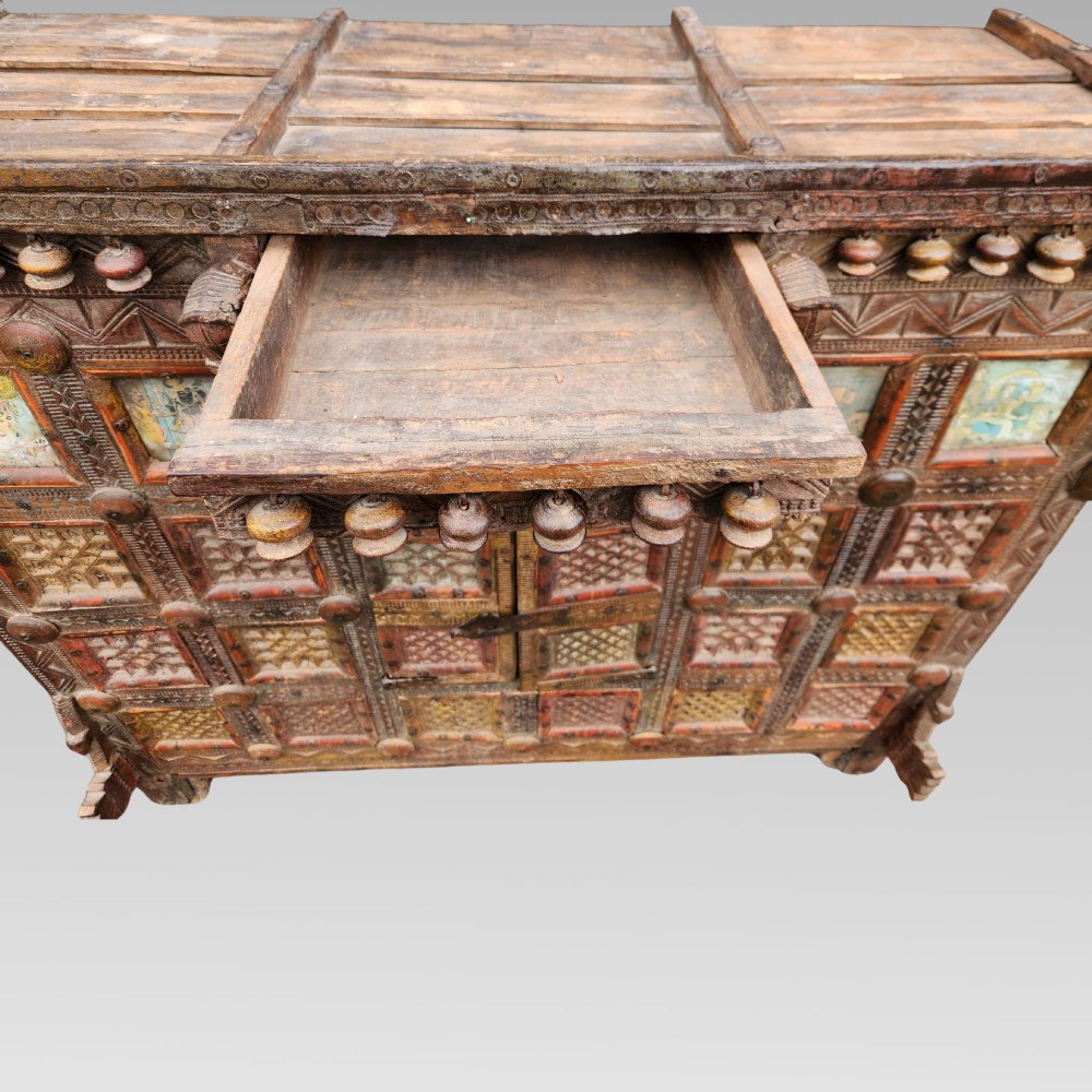 An intricately carved antique Indian teak Damchiya dowry chest featuring painted glass panels, horse head bracket carvings, metal studs, and small wooden wheels. View V