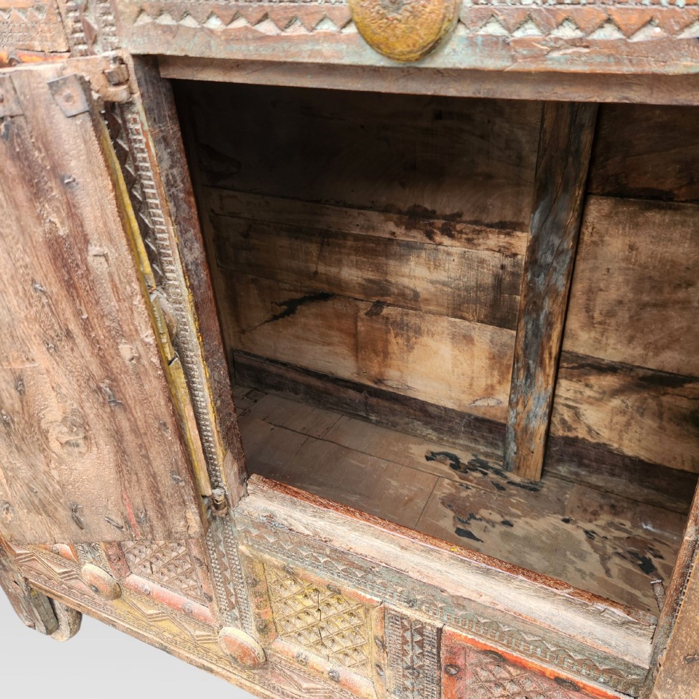 An intricately carved antique Indian teak Damchiya dowry chest featuring painted glass panels, horse head bracket carvings, metal studs, and small wooden wheels. View VI