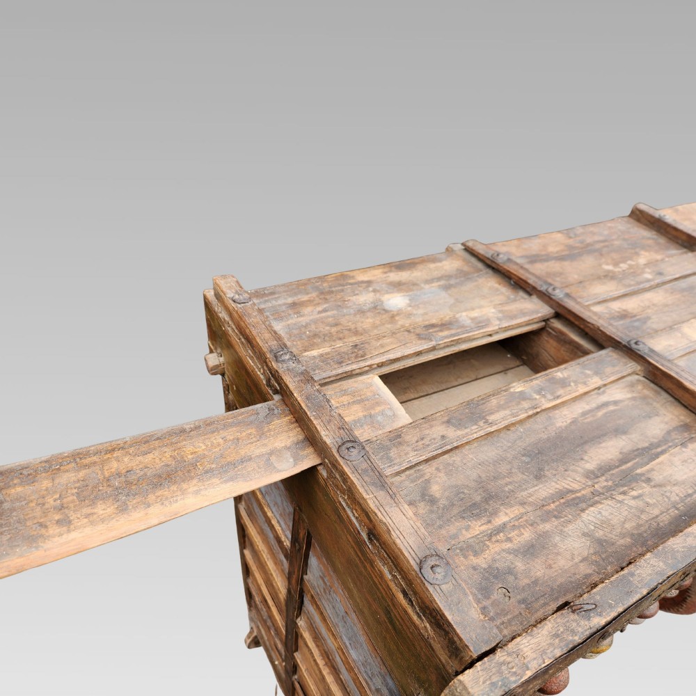 An intricately carved antique Indian teak Damchiya dowry chest featuring painted glass panels, horse head bracket carvings, metal studs, and small wooden wheels. View VII