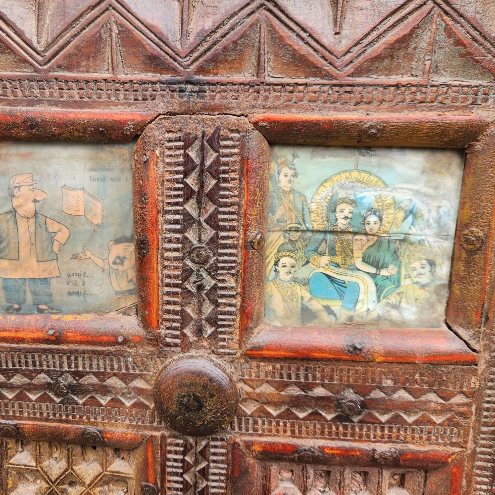 An intricately carved antique Indian teak Damchiya dowry chest featuring painted glass panels, horse head bracket carvings, metal studs, and small wooden wheels. View X