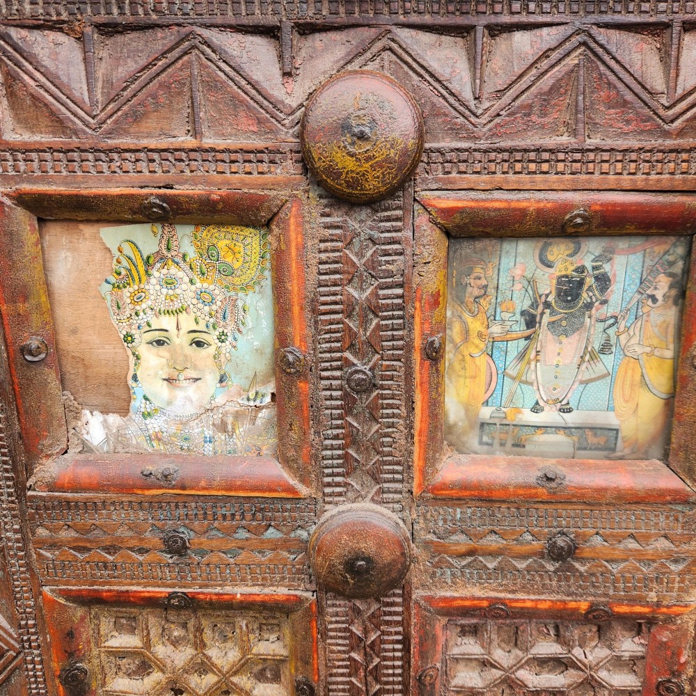 An intricately carved antique Indian teak Damchiya dowry chest featuring painted glass panels, horse head bracket carvings, metal studs, and small wooden wheels. View XI