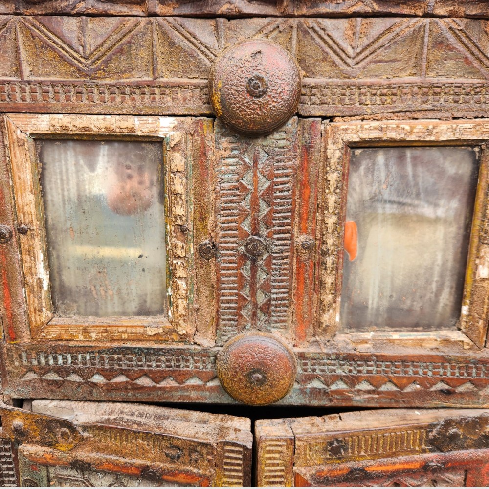 An intricately carved antique Indian teak Damchiya dowry chest featuring painted glass panels, horse head bracket carvings, metal studs, and small wooden wheels. View XII